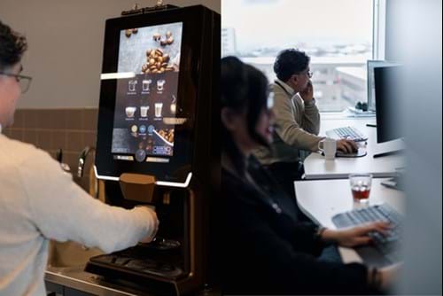 en kollage med to bilder av en mann som fyller en kaffekopp ved en maskin, og en mann som sitter ved en kontorpult og ser på en pc-skjerm.