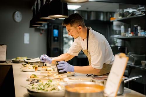en mann med brunt hår, briller, hvit kokkejakke, blått forkle og blå plasthansker som står på et arbeidskjøkken ved et hvitt bord og heller en spiseskje med chilisaus over en tallerken med en blomkålrett.